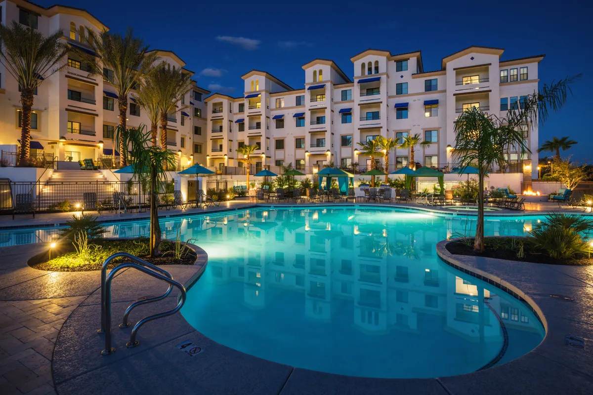An elegant exterior view featuring the swimming pool bordered by palm trees, stylish lounge chairs, and inviting canopy tents. The building’s facade and the pool deck exude an upscale, romantic vibe, perfect for sunset views or evening gatherings.