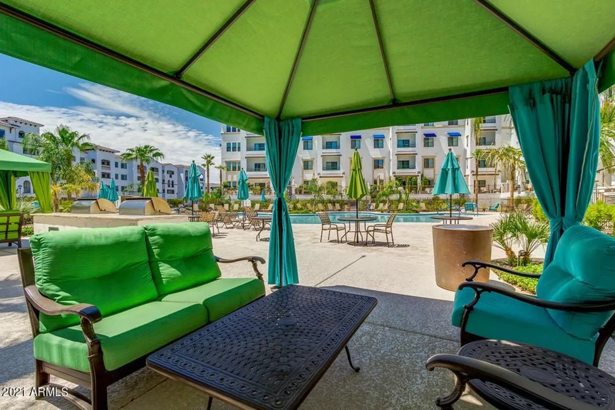 A lush pool area featuring a green cabana, numerous lounge chairs arranged around the pool, vibrant potted plants, and palm trees. The scene exudes a relaxed atmosphere ideal for socializing, sunbathing, or taking a dip. The cabana and greenery create a resort-like ambiance.