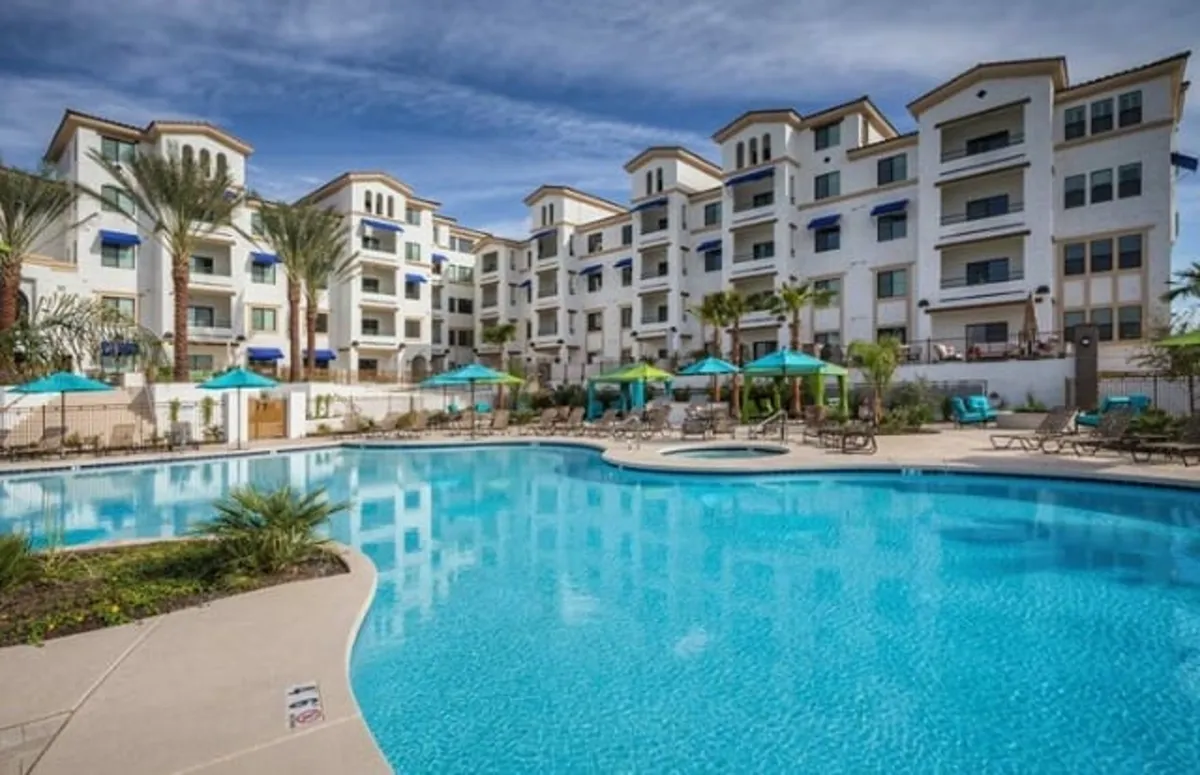 A sparkling swimming pool lined with palm trees and lounge chairs, set against the backdrop of a residential building. The inviting water, umbrellas, and seating create a cheerful, relaxed environment for swimming, sunbathing, or enjoying time with friends.
