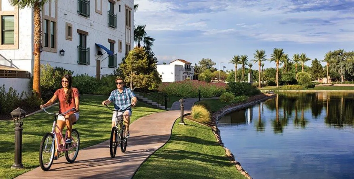 Two cyclists enjoy a ride along a scenic pathway bordered by palm trees and a shimmering lake, with the residences and blue sky visible in the background. The setting is lively, active, and suggests easy access to outdoor recreation.