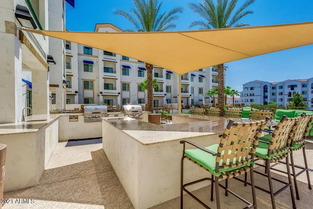 A vibrant outdoor grill and bar area, complete with ample seating, surrounded by palm trees and shaded by a modern sail canopy. The atmosphere is lively and welcoming, perfect for socializing, dining outdoors, or relaxing with friends and neighbors.