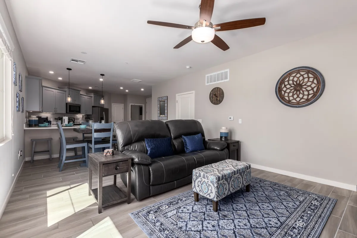 A stylish interior living room featuring a comfortable black leather sofa, a decorative ottoman with a blue pattern, a plush area rug, and a ceiling fan with a light fixture. The space is arranged for both relaxation and entertaining, showcasing a modern yet inviting aesthetic.