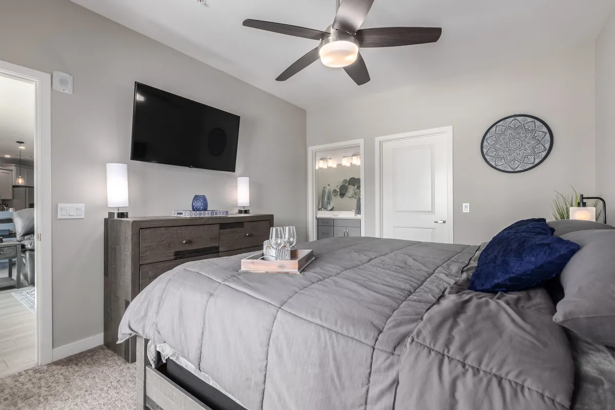 A sophisticated bedroom scene with a bed dressed in gray luxury linens, a dark wood dresser, a TV mounted on the wall, decorative wall art, and a nightstand with glasses and a tray. The mood is serene and cozy, suitable for quiet evenings and mornings in bed.