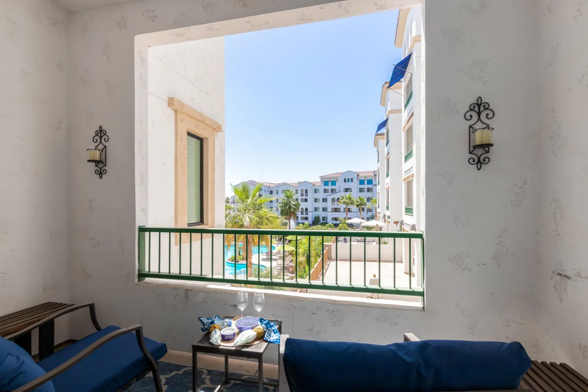 An elevated balcony or patio overlooking the sparkling resort pool, with stylish blue seating, decorative wall sconces, and a setup featuring drinks and snacks. The tranquil setting suggests a perfect spot for relaxing, socializing, or enjoying morning coffee with a view of the amenities below.