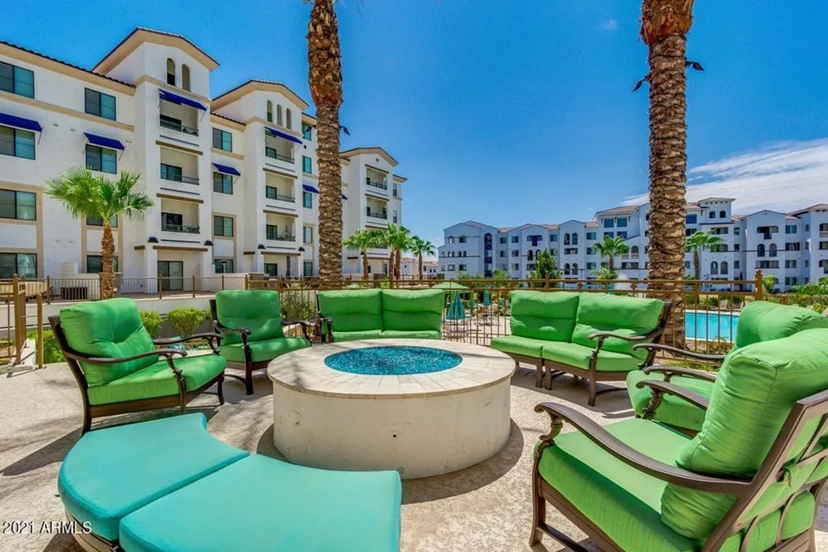 A stylish outdoor lounge space featuring a vibrant green seating area, a glowing fire pit, and palm trees. The pool shimmers in the background, setting the scene for evening gatherings and socializing with neighbors.