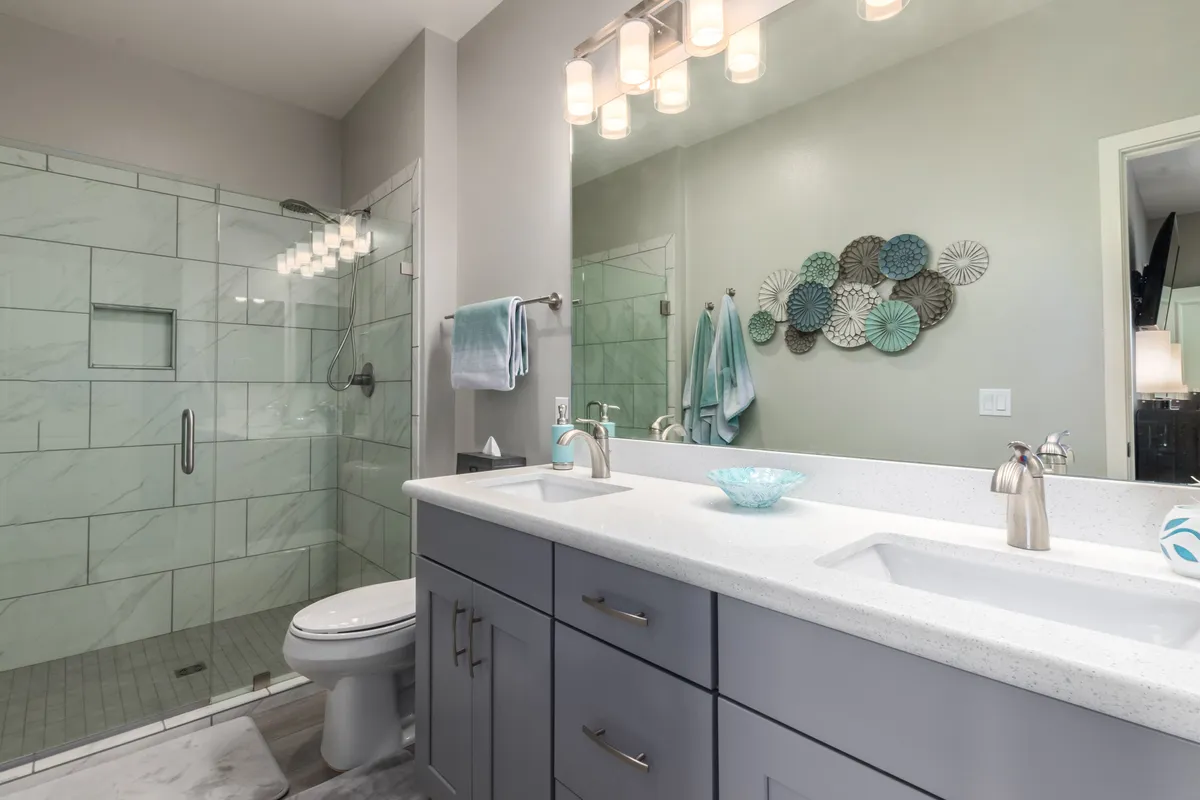 A bright and spa-inspired ensuite bathroom with dual granite sinks, a glass shower, and decorative wall art. The room is fresh and elegantly designed, reflecting a serene atmosphere ideal for self-care routines.