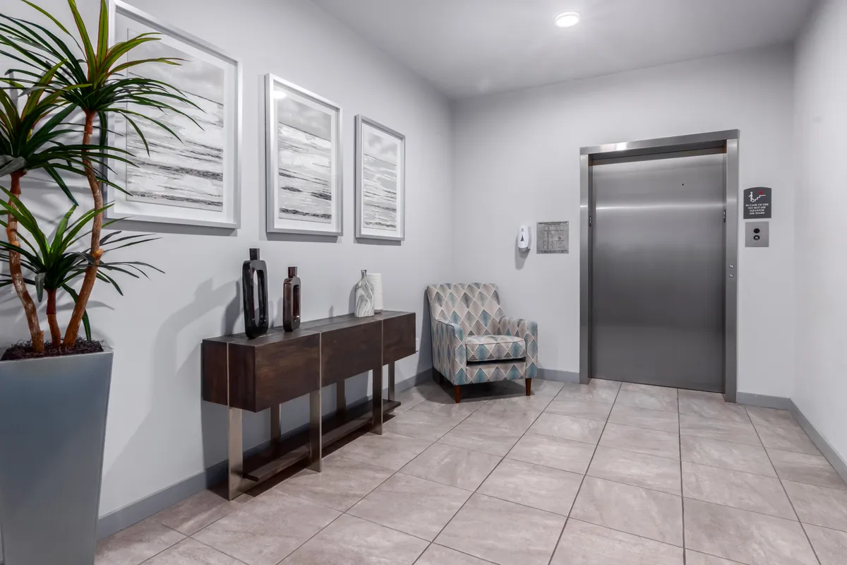 A calm, inviting common area in the building, featuring an elevator, artwork, a console table, and a comfortable chair with a potted plant. The space evokes a sense of security and community.