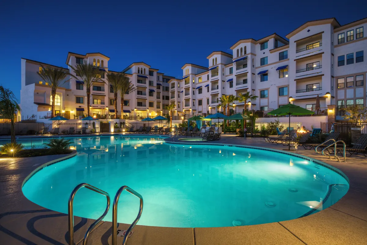 A sparkling swimming pool surrounded by modern resort buildings, tall palm trees, rows of lounge chairs, and inviting umbrellas. The space is lively yet relaxing, with guests swimming and others unwinding in shaded cabanas or sunbathing beside the water.